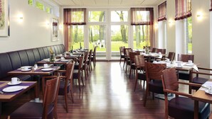 Un restaurante con varias mesas y sillas, amplias ventanas con cortinas, y vista a un área verde al aire libre.