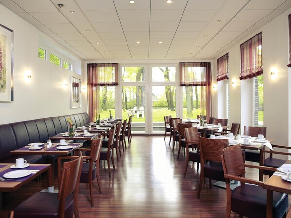 Een restaurant met meerdere tafels en stoelen, grote ramen met gordijnen en uitzicht op een groen buitengebied.