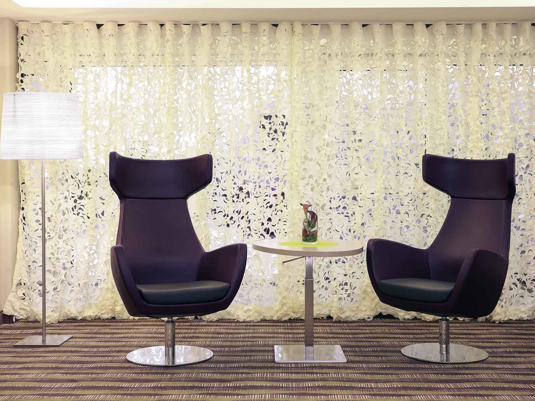 Two modern chairs with a round table between them, a decorative vase on the table, and a patterned curtain in the background.