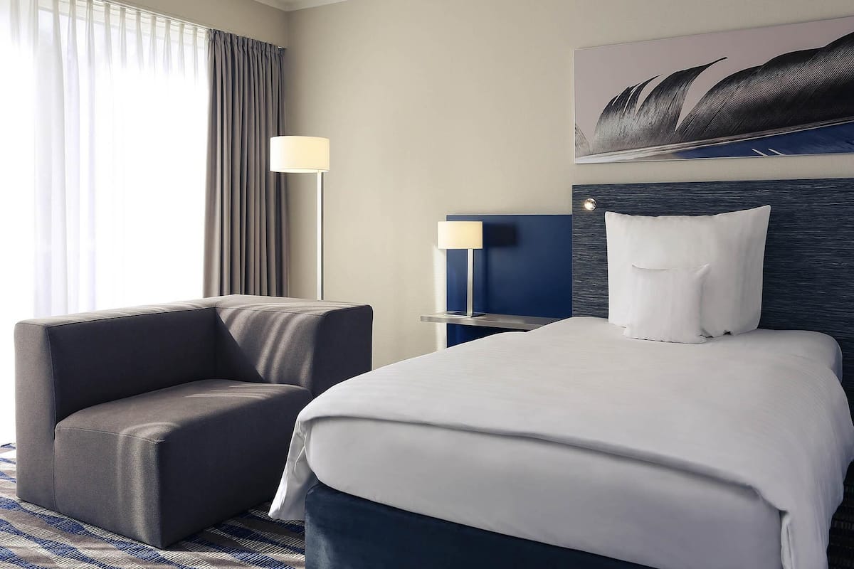 A hotel room with a bed, a chair, a nightstand, a lamp, and a painting on the wall.