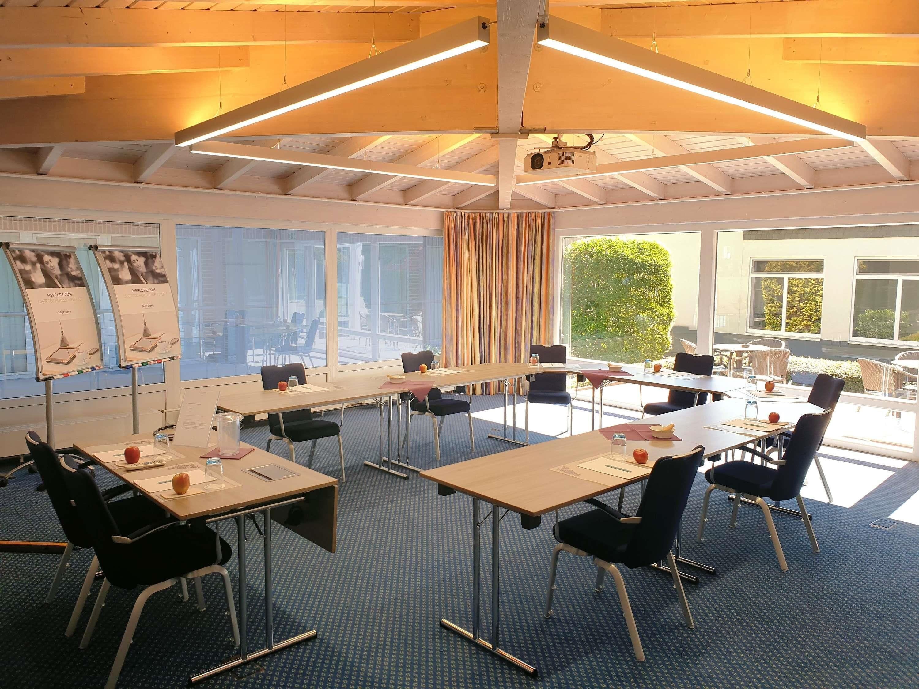 Un salón de conferencias con mesas, sillas, un proyector y ventanas amplias.