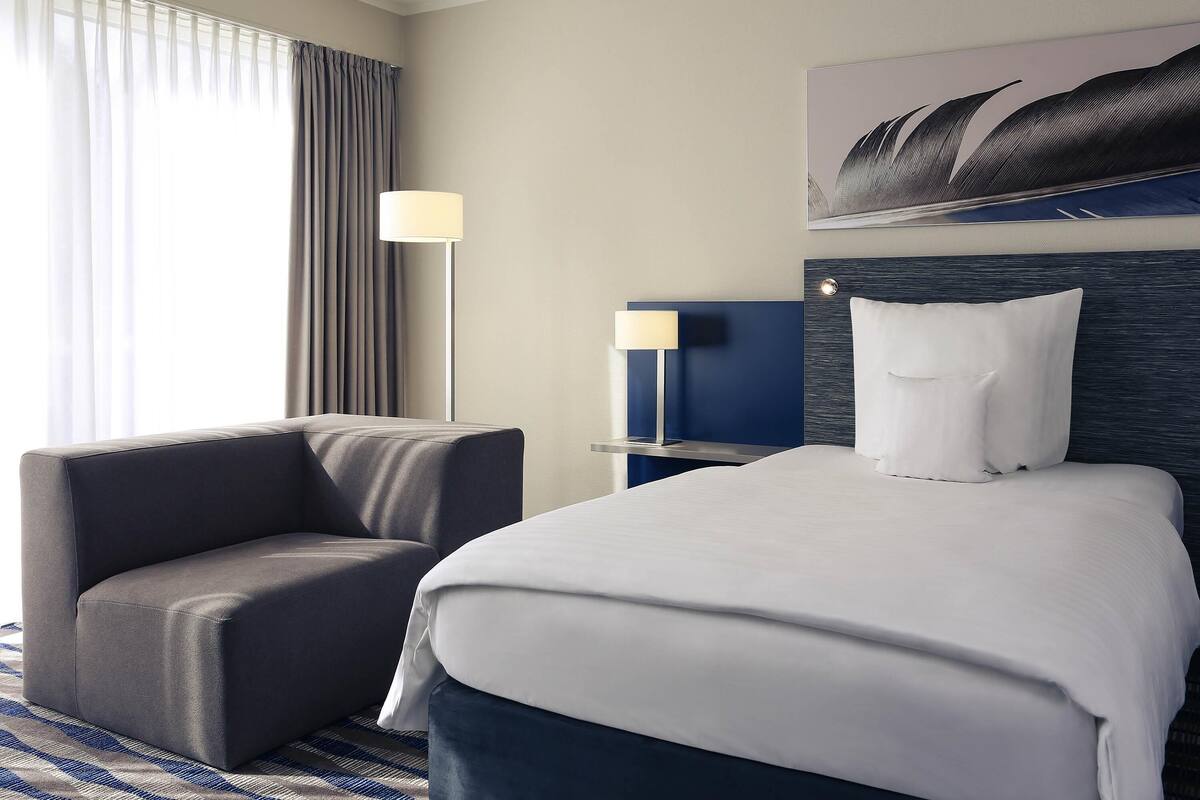 Quarto de hotel com cama, cadeira, mesa de cabeceira, luminária e um quadro na parede.