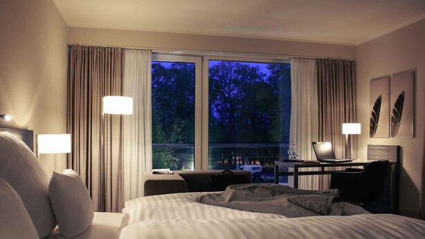 Habitación de hotel con cama, escritorio y computadora portátil, y vista a los árboles a través de la ventana.