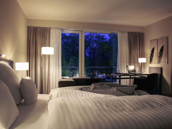 Habitación de hotel con cama, escritorio y computadora portátil, y vista a los árboles a través de la ventana.
