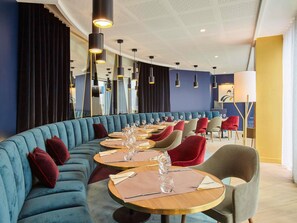 Ein modernes Restaurant mit einer geschwungenen Sitzgruppe in Blau, runden Tischen mit Holzbeschlägen und schwarzen Pendelleuchten.