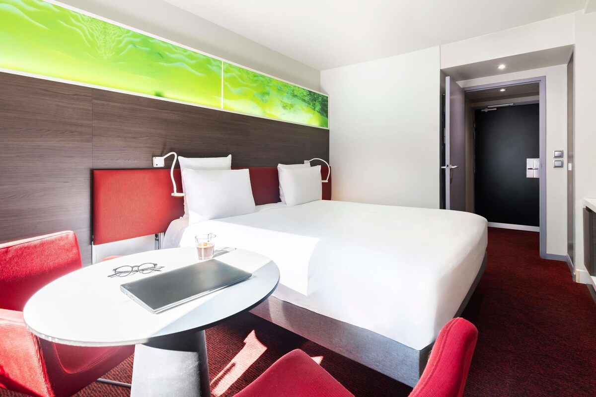 A hotel room with a bed, a round table, red chairs, a wall-mounted lamp, and a door leading to another room.