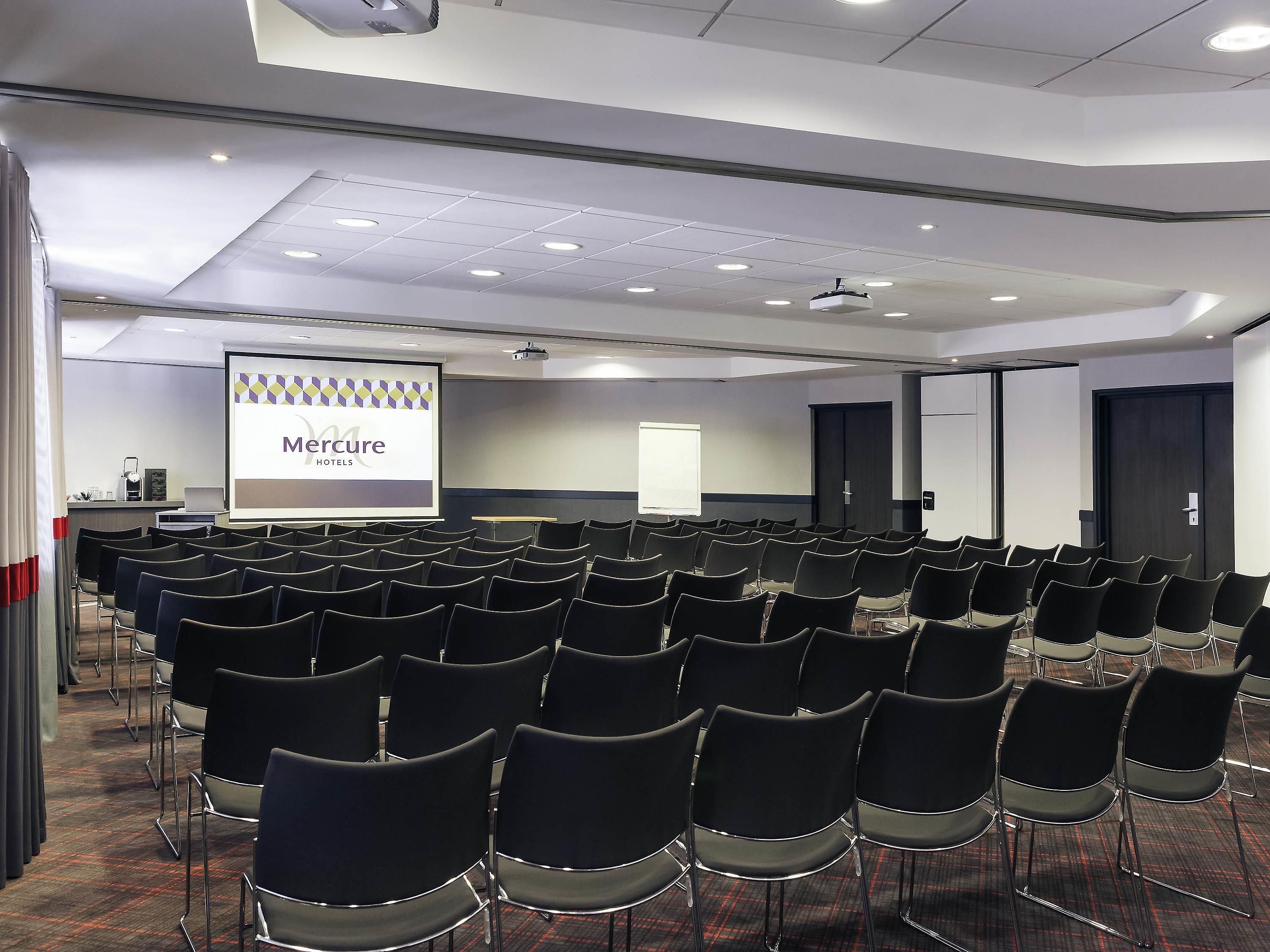 Una sala conferenze con file di sedie, uno schermo per proiezioni con una presentazione e una porta sullo sfondo.