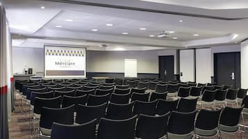 Ein Konferenzraum mit Reihen von Stühlen, einer Projektionsleinwand, auf der eine Präsentation zu sehen ist, und einer Tür im Hintergrund.