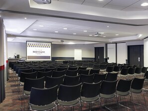 A conference room with rows of chairs, a projector screen displaying a presentation, and a door in the background. - Mercure Paris La Défense Grande Arche (Nanterre)