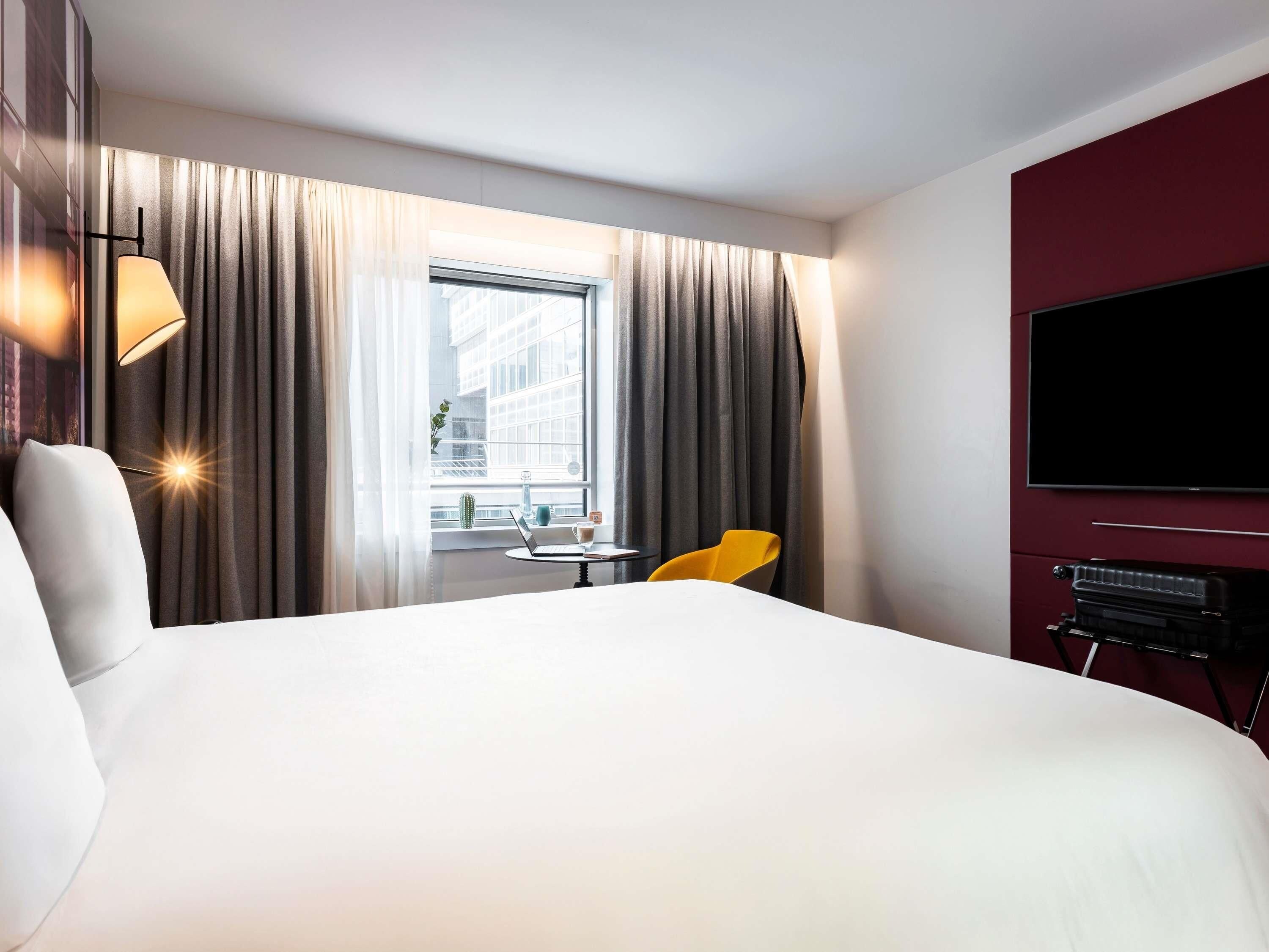 A hotel room with a large bed, a small table with a laptop, a chair, a TV, and curtains.