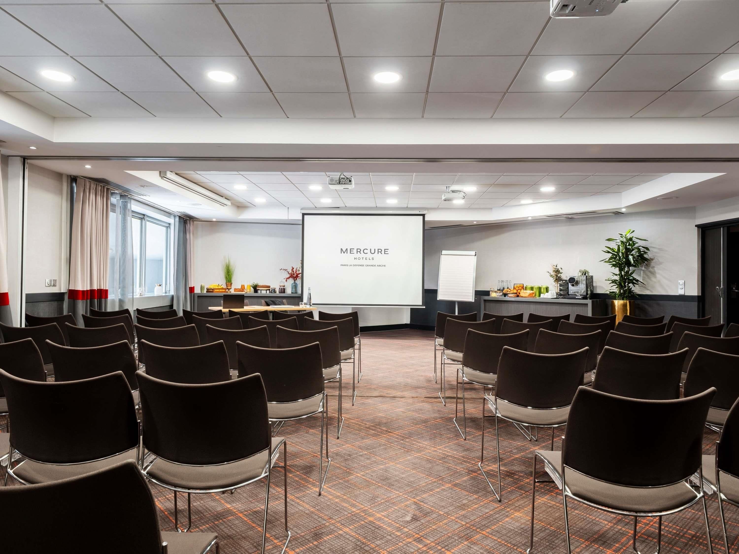 Un salón de conferencias con filas de sillas, una pantalla de presentación, una pizarra y un mostrador de recepción con plantas y refrigerios.