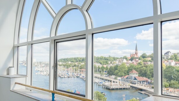 Uitzicht door een groot raam met houten leuning, op een jachthaven met boten, een kerk en een stad met groen.