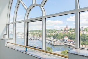 Blick durch ein großes Fenster mit Holzgeländer auf einen Yachthafen mit Booten, eine Kirche und eine grüne Stadt.