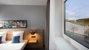 Een hotelkamer met een bed, een nachtkastje, een wandlamp en een groot raam met uitzicht op een rivier en gebouwen.