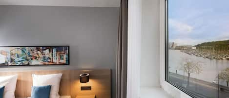 A hotel room with a bed, a nightstand, a wall-mounted lamp, and a large window with a view of a river and buildings.
