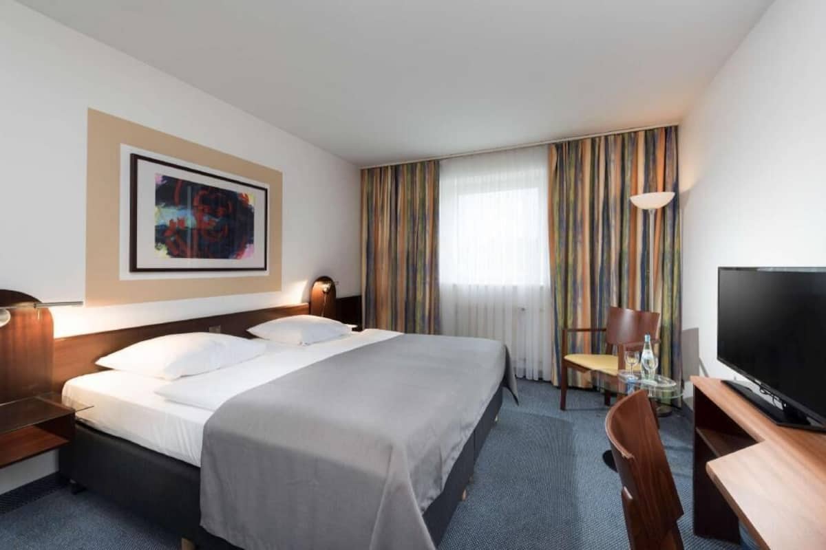 A hotel room with a bed, a desk with a TV, a chair, a lamp, a window with curtains, and a painting on the wall.
