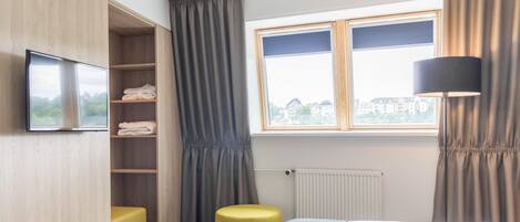 A hotel room with a bed, a TV, a window with curtains, a radiator, and a yellow ottoman.
