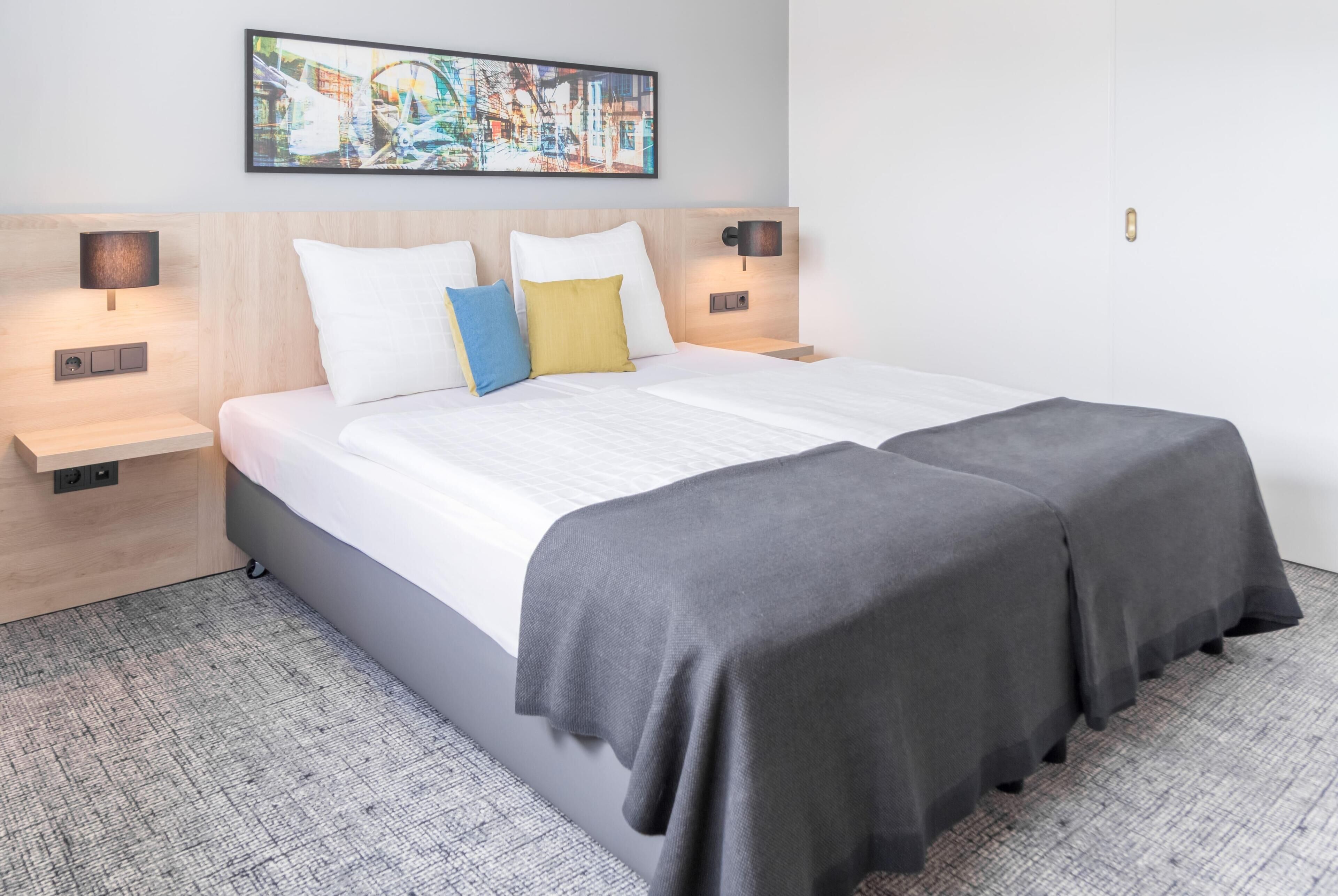 Habitación de hotel con una cama grande, dos mesitas de noche, una lámpara de pared, un cuadro y suelo alfombrado.