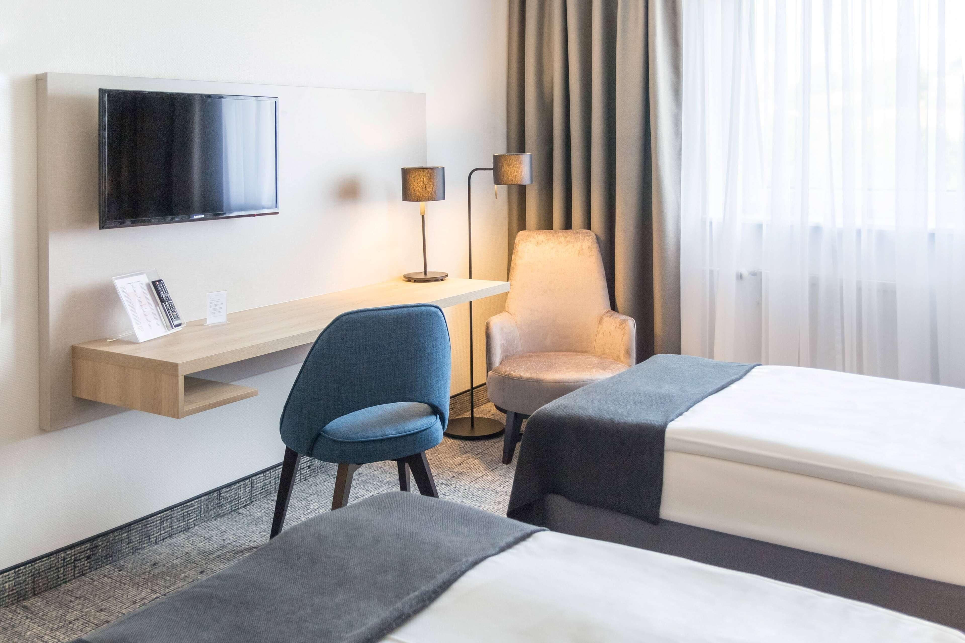 Habitación de hotel con una televisión montada en la pared, un escritorio con una lámpara, dos sillas, una cama y una ventana con cortinas.
