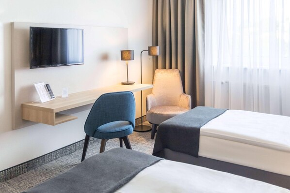 A hotel room with a TV mounted on the wall, a desk with a lamp, two chairs, a bed, and a window with curtains.