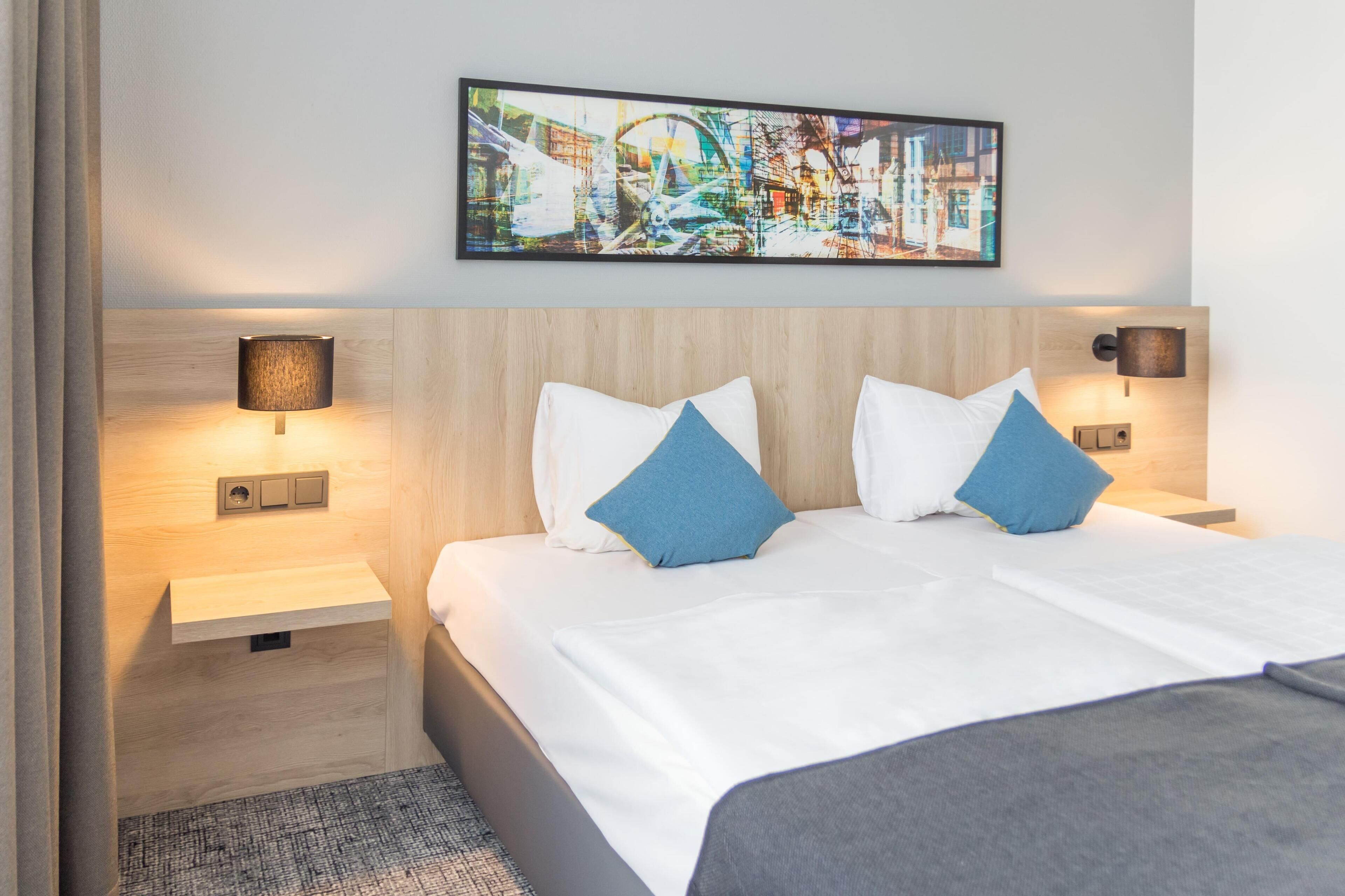 A hotel room with a bed, two bedside tables, wall-mounted lamps, a wooden headboard, and a framed picture above the bed.