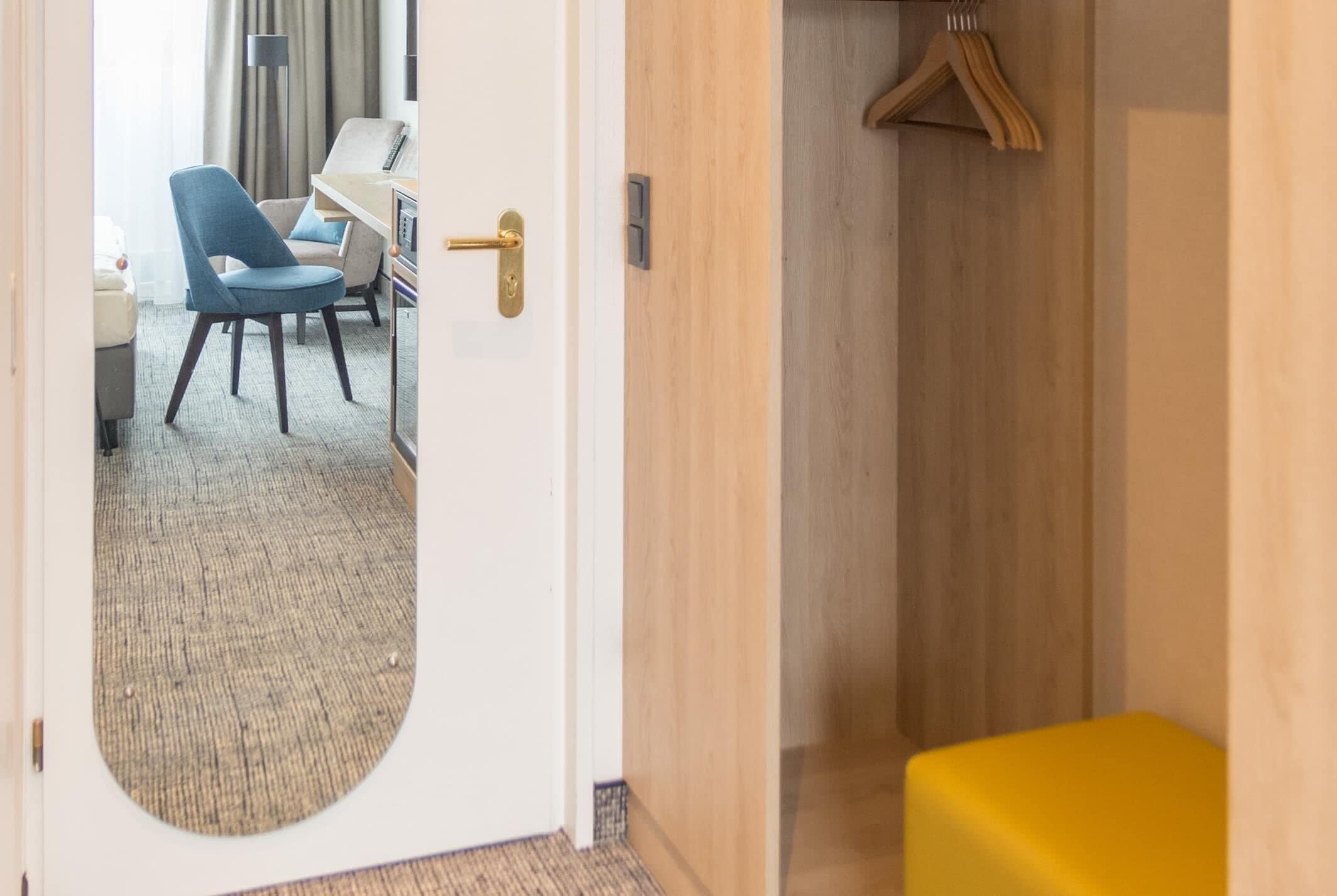 Una habitación de hotel con un espejo redondo, una silla azul, un banco de madera y un otoman amarillo.