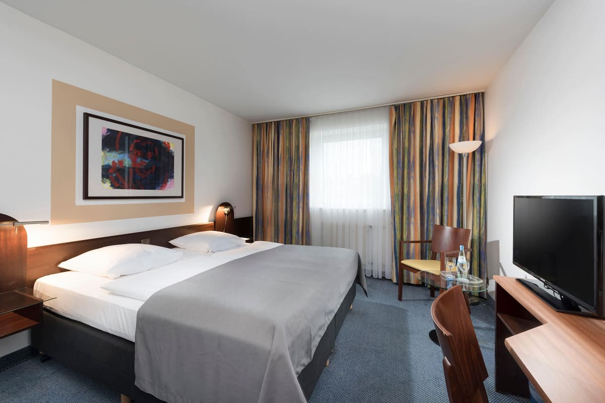 Habitación de hotel con cama, escritorio con televisor, una silla, una mesa con vaso y ventana con cortinas.