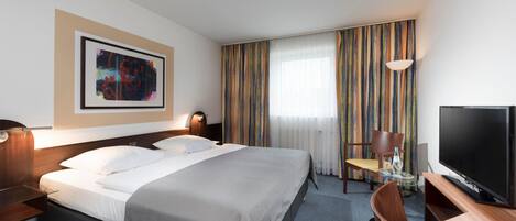 Habitación de hotel con cama, escritorio con televisor, una silla, una mesa con vaso y ventana con cortinas.