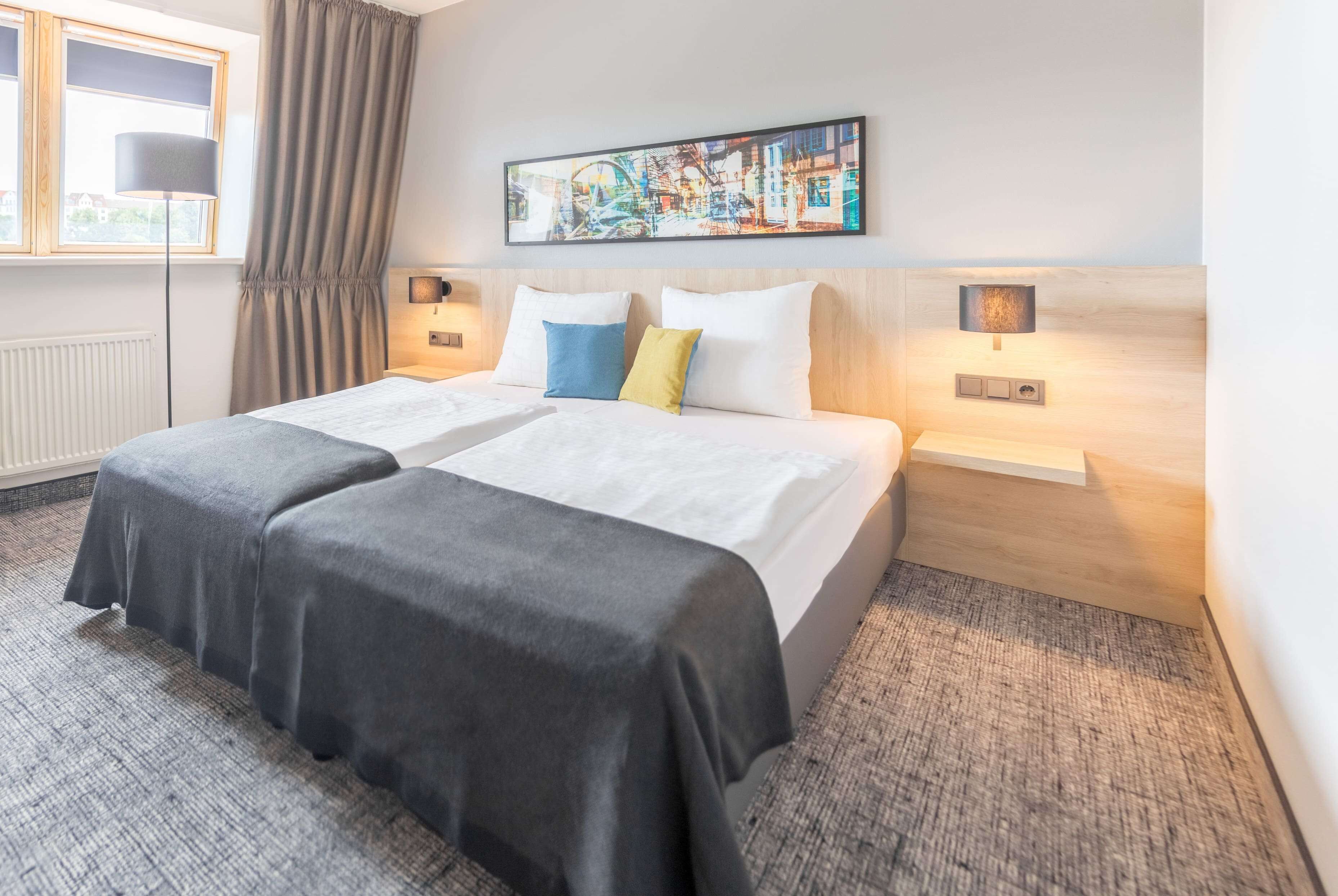 A double bed with a grey bedspread, two pillows in blue and yellow, and a headboard with a cityscape painting. There are two bedside tables with lamps, a window with curtains, and a radiator.