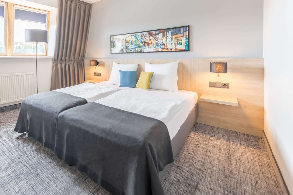 Cama doble con colcha gris, dos almohadas en azul y amarillo, y cabecera con pintura de un paisaje urbano. Hay dos mesitas de noche con lámparas, una ventana con cortinas y un radiador.