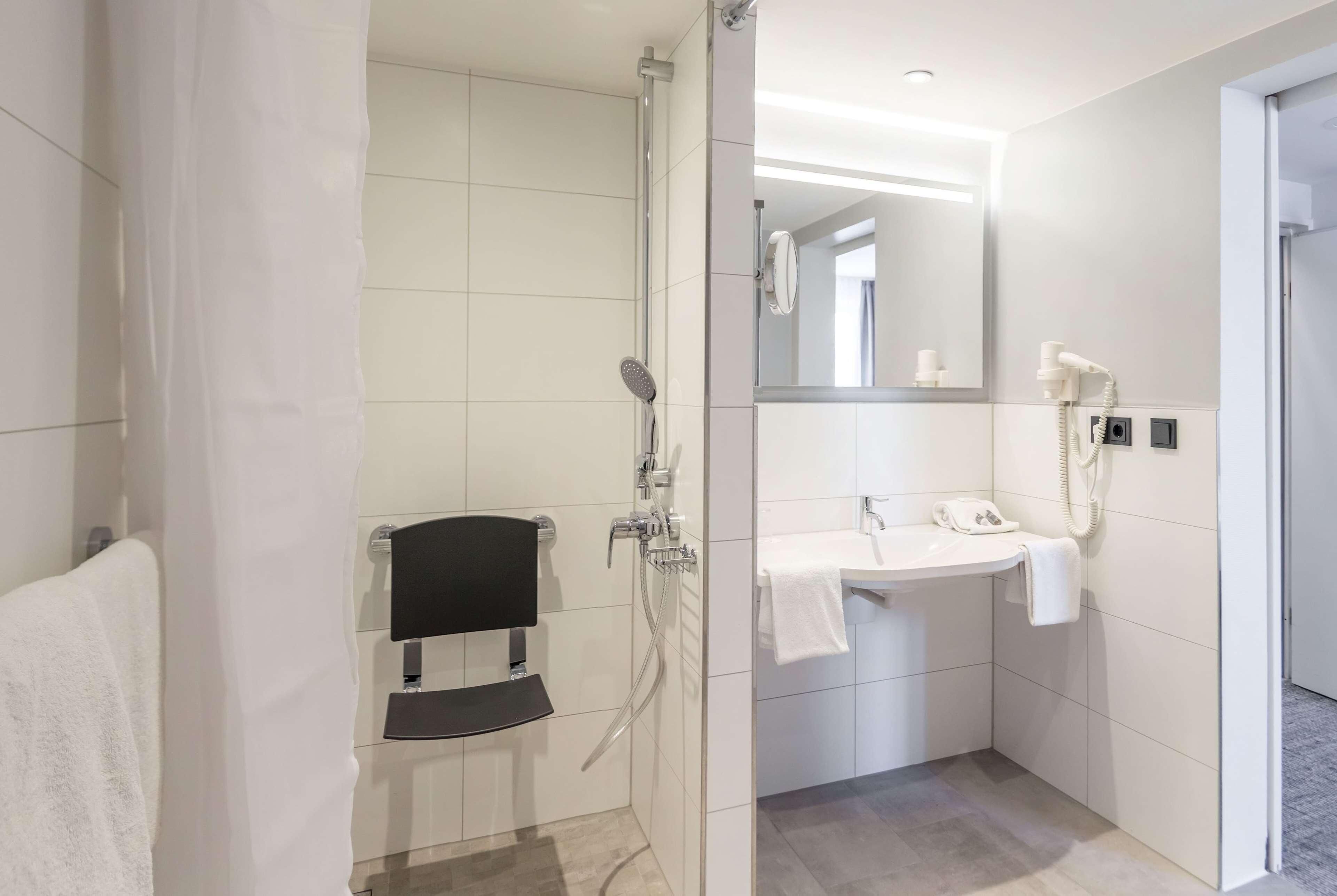 Un baño moderno con ducha envuelta en vidrio, toallero negro, lavamanos blanco con espejo y toalla blanca.