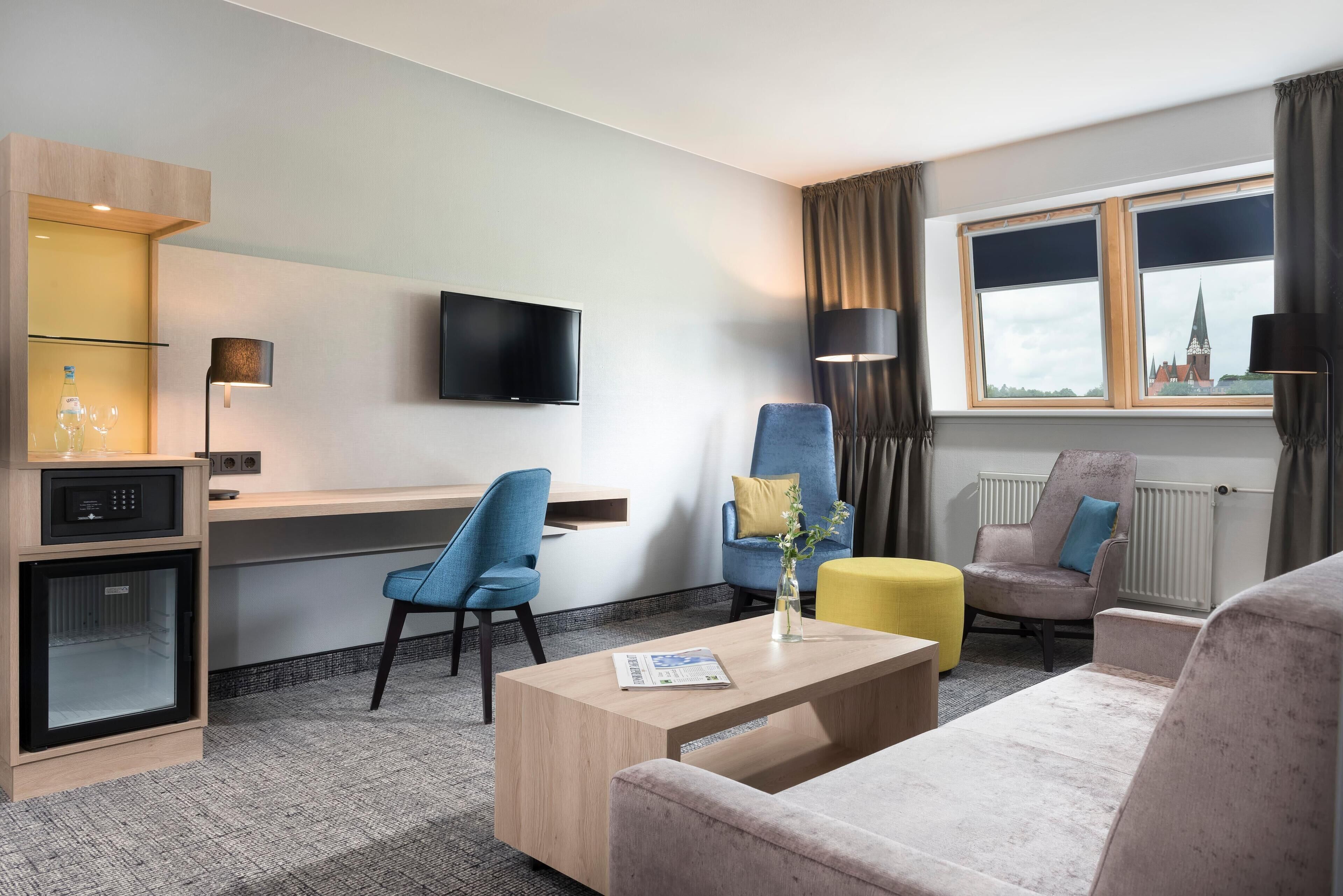 Habitación de hotel con televisión de pantalla plana, escritorio, dos sillas, sofá, mesa de centro y ventana con vista a un edificio.