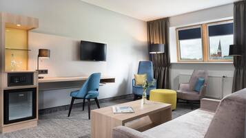 Habitación de hotel con televisión de pantalla plana, escritorio, dos sillas, sofá, mesa de centro y ventana con vista a un edificio.