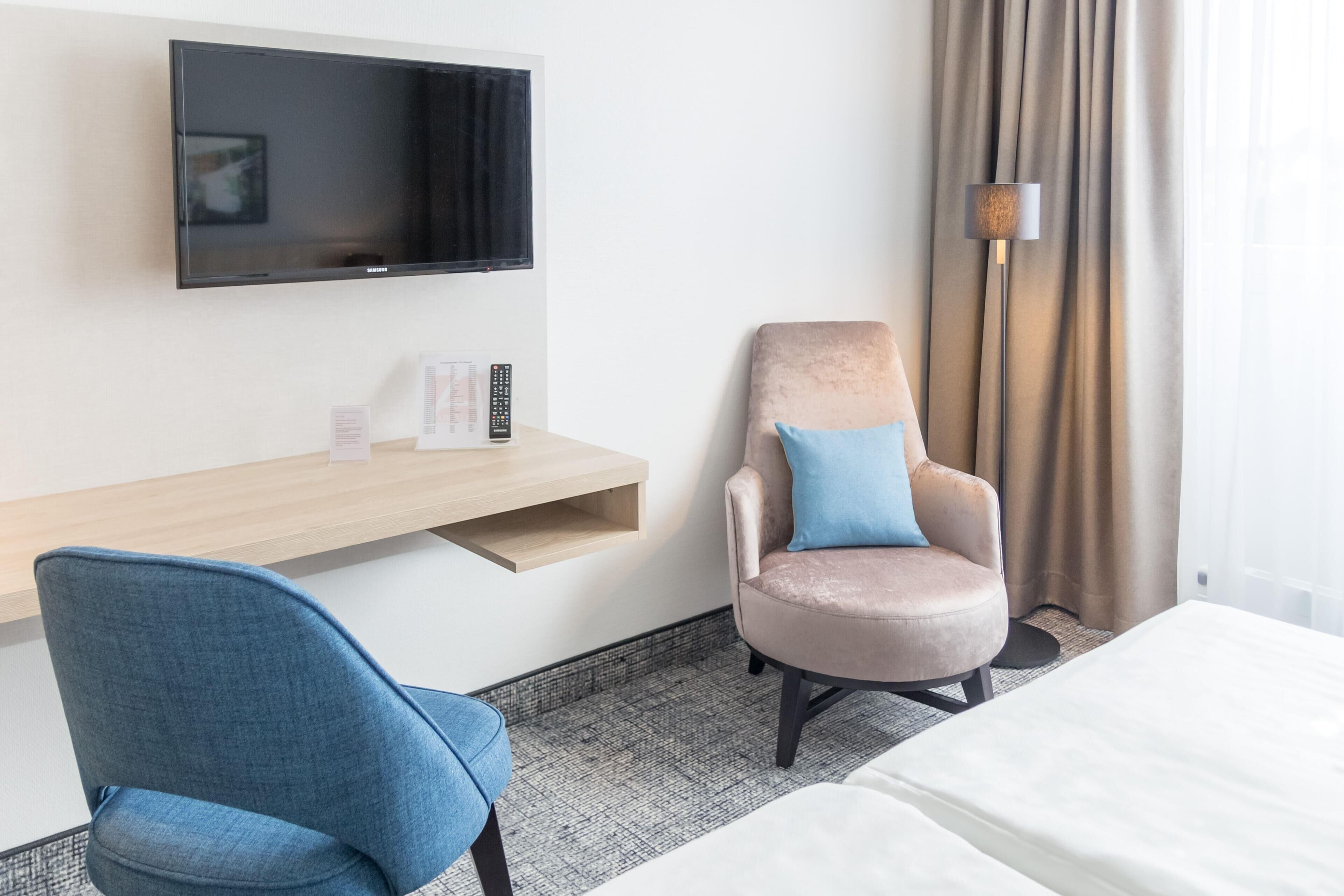 Habitación de hotel con una televisión de pantalla plana, un escritorio con control remoto, una silla azul, un sillón color beige con almohada azul, y una cama.