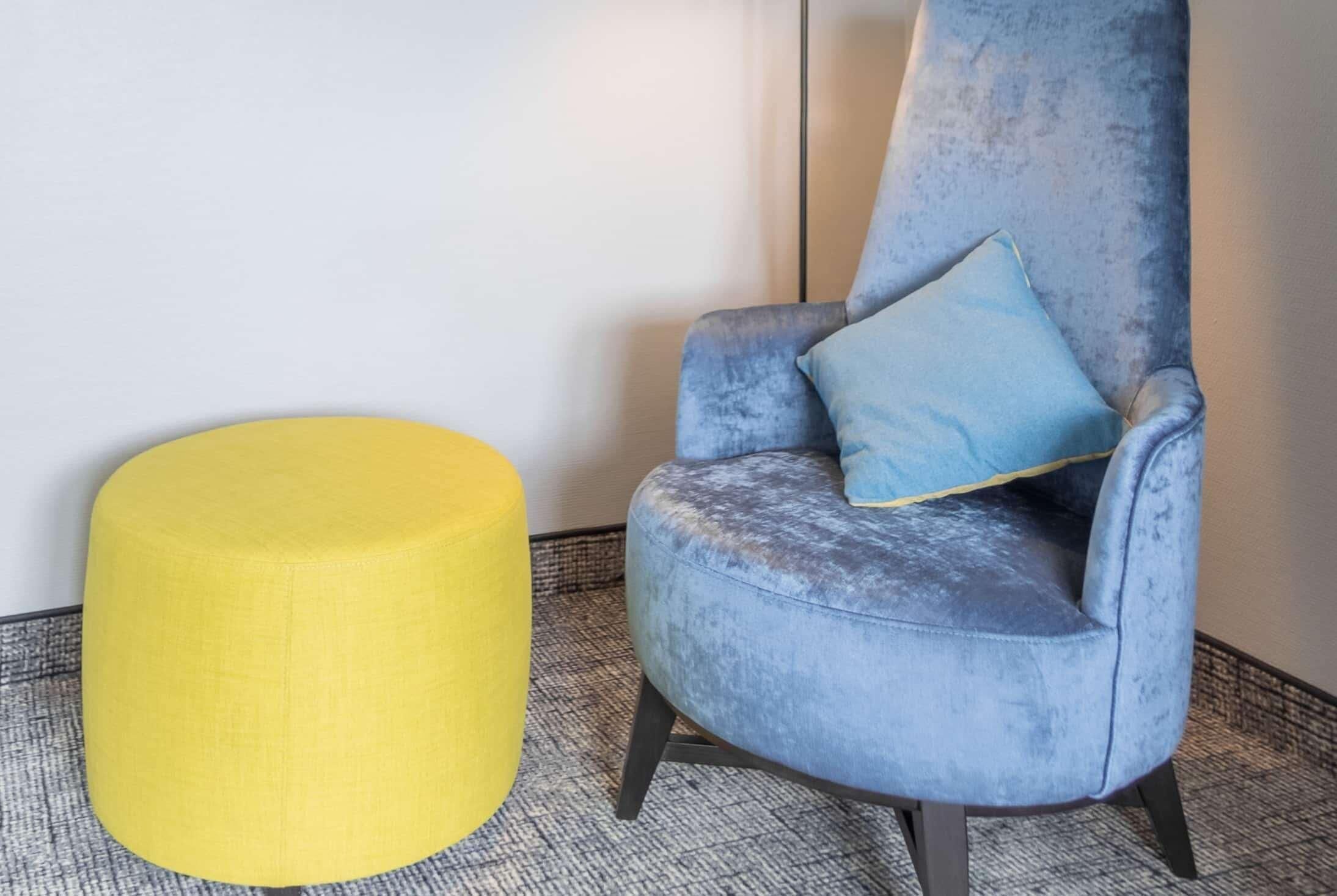 Une chaise avec un coussin et un pouf jaune dans une pièce dont le sol est recouvert d’un tapis à motifs.