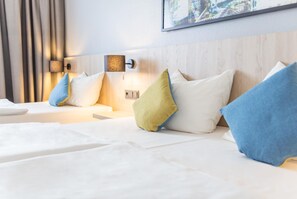 A hotel room with two beds, each with white linens and multiple pillows in various colors. There is a wall-mounted lamp and a painting above the beds.