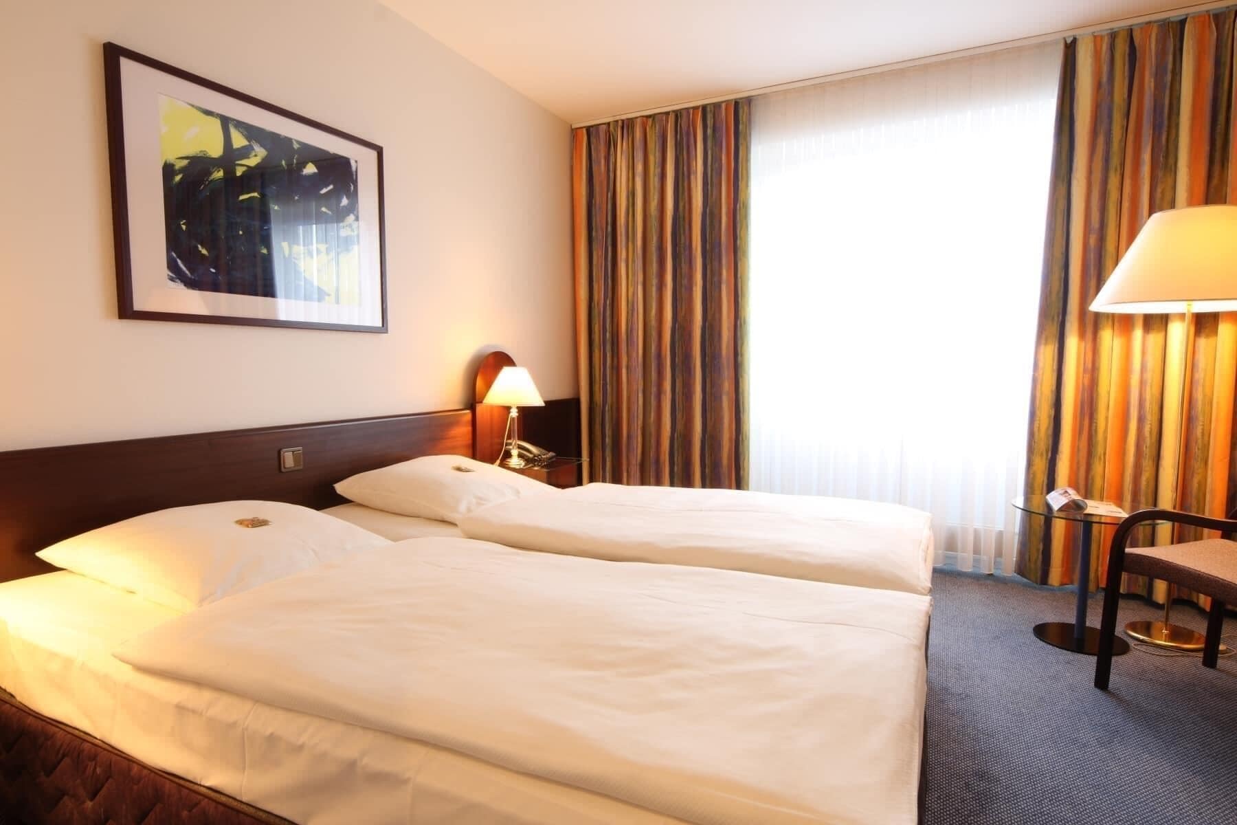 A hotel room with two beds, a desk, a chair, a lamp, and a painting on the wall.