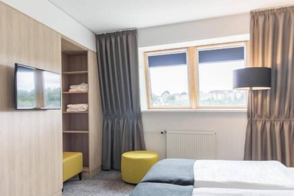 Una habitación de hotel con cama, televisor, ventana con cortinas, radiador y armario.