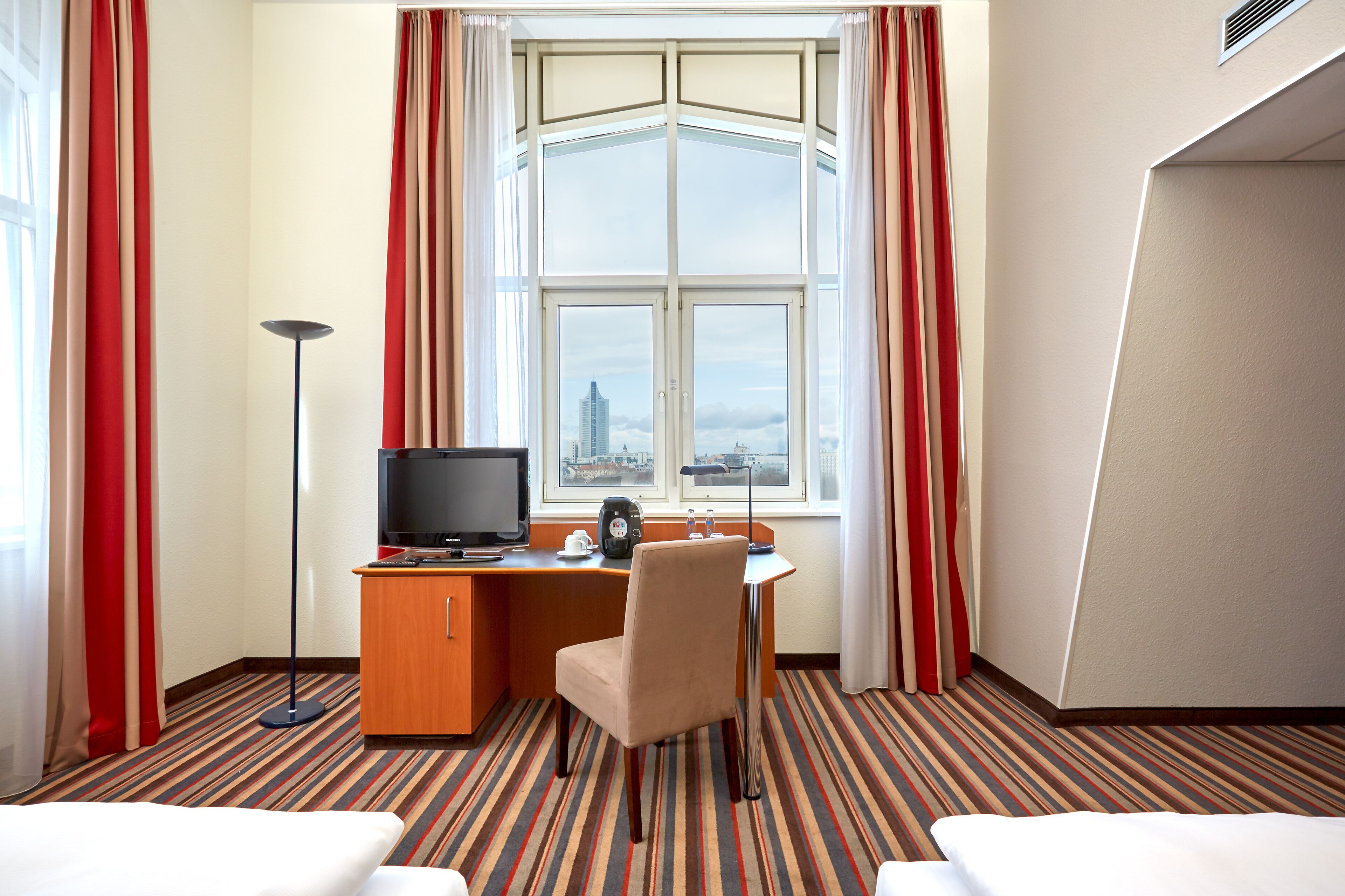 Quarto de hotel com cama, escrivaninha com computador e cafeteira, uma cadeira e uma janela grande com cortinas.