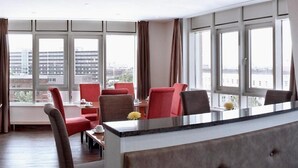 Ein modernes Hotelzimmer mit großem Fenster, einer Essecke mit roten Stühlen und einer zentralen Insellounge mit Blumenvase.