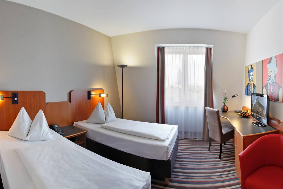 Habitación de hotel con dos camas, un escritorio, una silla, una lámpara y obras de arte en la pared.