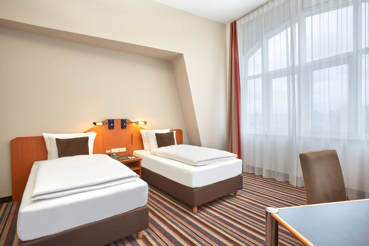 Una habitación de hotel con dos camas, una mesita de noche, una silla y un ventanal grande con cortinas.