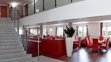 Um saguão de hotel com escadaria, balcão de recepção e área de refeições com cadeiras e mesas vermelhas.