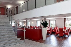 A hotel lobby with a staircase, a reception desk, and a dining area with red chairs and tables. - H+ Hotel Leipzig (Leipzig)