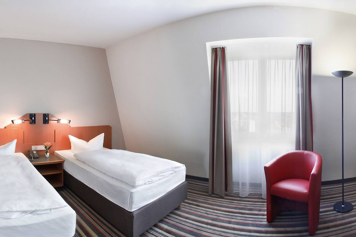 Habitación de hotel con dos camas, una mesita de noche, una silla roja, una lámpara de pie y un ventanal grande con cortinas.