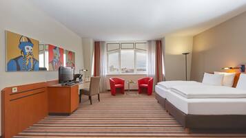 Ein Hotelzimmer mit einem Bett, einem Schreibtisch mit Fernseher, zwei roten Stühlen, einem kleinen Tisch und einem Fenster mit Vorhängen.