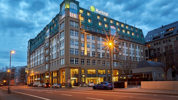 Ein mehrstöckiges Hotel mit einem gut sichtbaren H+Hotel-Schild, großen Fenstern und einer gut beleuchteten Fassade.