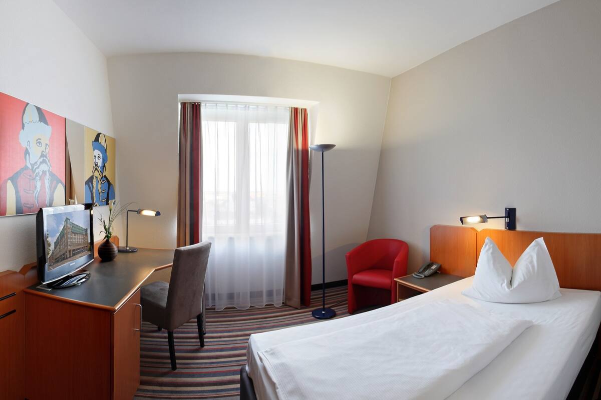 Habitación de hotel con cama, escritorio, silla, obras de arte y ventana con cortinas.