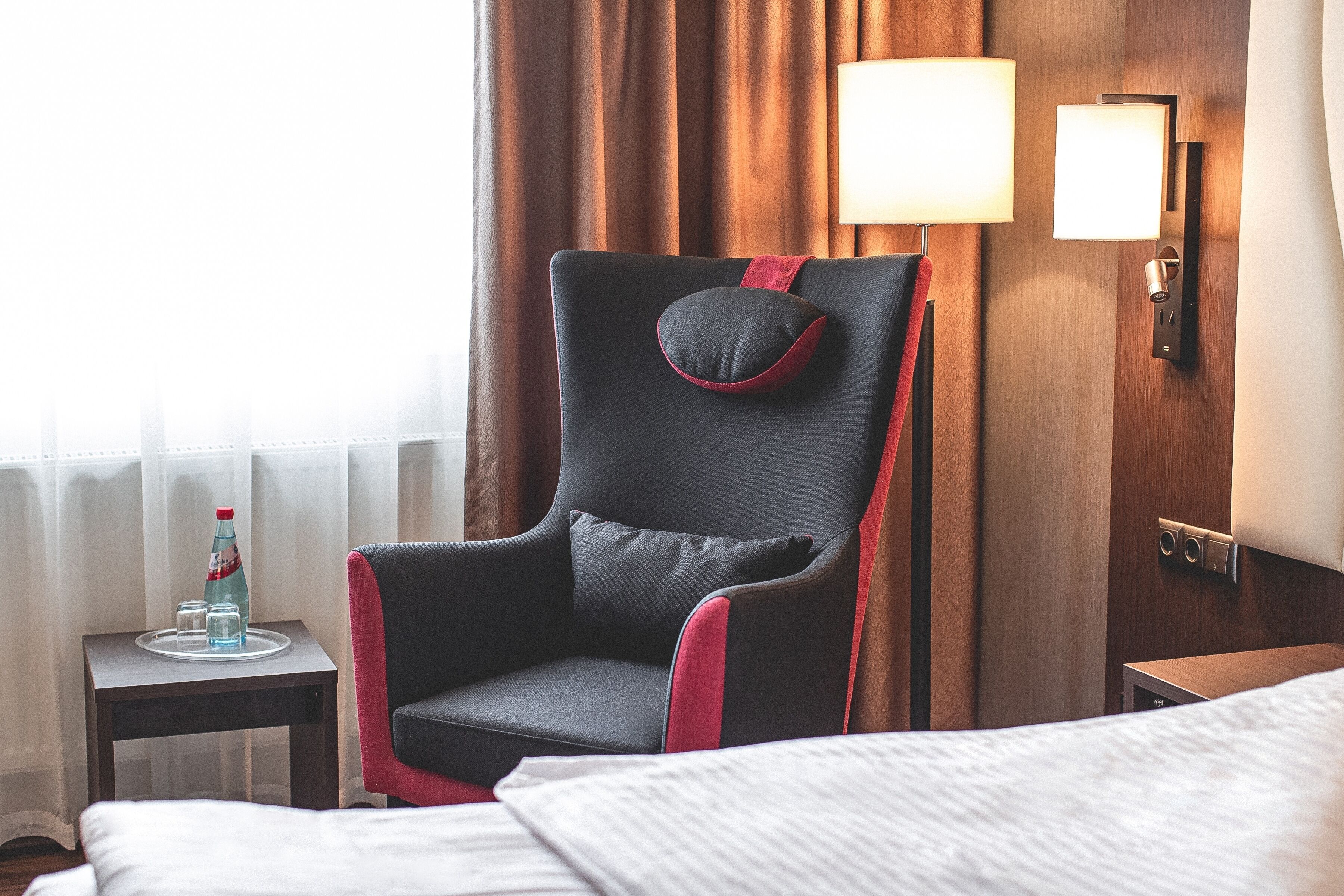 A hotel room with a bed, a chair, a nightstand, a lamp, and a window with curtains.