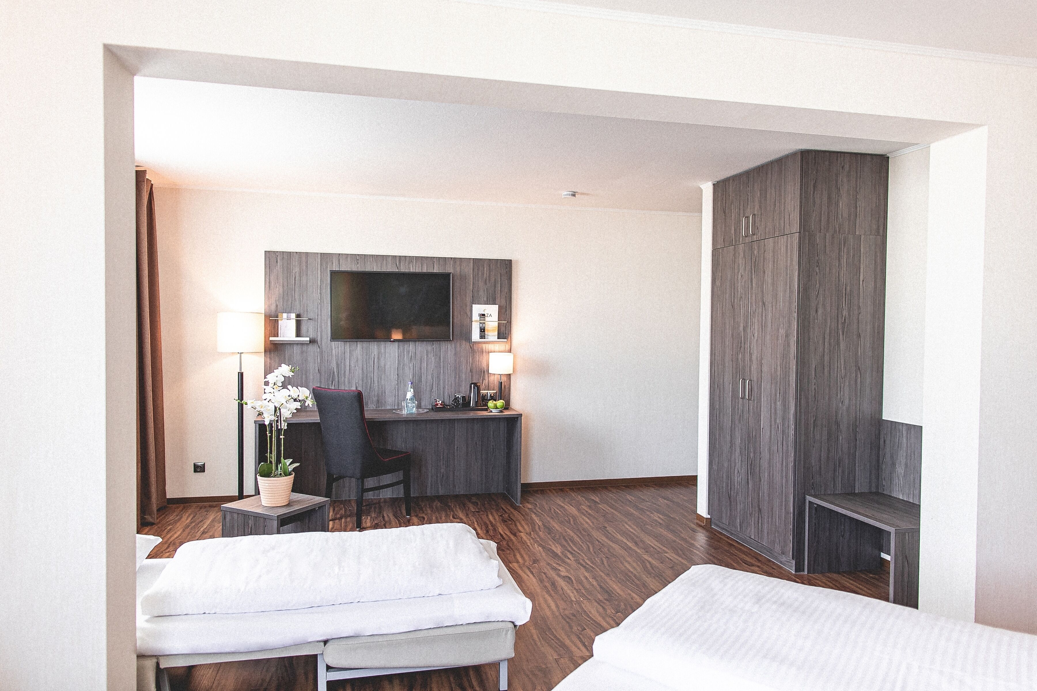 Habitación de hotel con dos camas, un escritorio con silla, televisor montado en la pared y un armario.